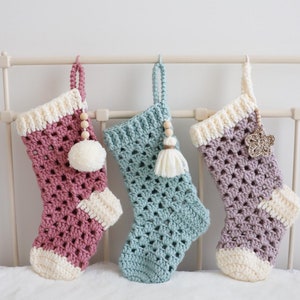 Puede incluir: Tres calcetines de Navidad de ganchillo colgados de un marco de cama de metal blanco. Los calcetines son rosa, azul verdoso y morado con ribetes blancos. Cada calcetín tiene una cuenta de madera y un adorno diferente: un pompón blanco, un borla azul verdoso y una estrella dorada.