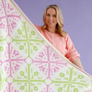 Peut inclure: Une couverture au crochet faite à la main avec un motif géométrique en blanc, rose et vert. La couverture présente un motif floral répétitif et est tenue sur un fond violet. La couverture est un article de décoration intérieure confortable.