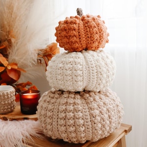 May include: A stack of three crocheted pumpkins in varying sizes and colors, featuring a textured bobble stitch pattern. The top pumpkin is burnt orange, the middle is cream, and the bottom is beige. Each pumpkin has a small brown stem on top.