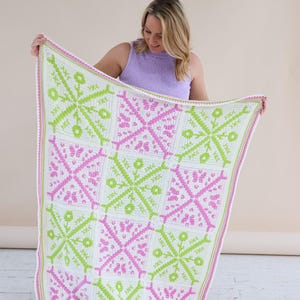 Puede incluir: Una manta de ganchillo hecha a mano con un patr&oacute;n geom&eacute;trico repetido en blanco, verde claro y rosa. La manta se sostiene, mostrando el intrincado dise&ntilde;o y la combinaci&oacute;n de colores. Los bordes est&aacute;n acabados con un borde rosa.