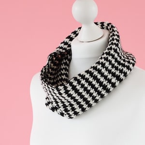 May include: A black and white houndstooth pattern infinity scarf displayed on a white mannequin against a pink background. The scarf is a continuous loop, designed to be worn around the neck. The fabric appears to be knitted or woven.