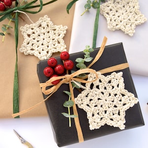 May include: Three gifts wrapped in brown paper, white paper, and black paper. Each gift is decorated with a crocheted snowflake ornament and red berries.