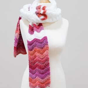 May include: A handmade white crochet scarf with a chevron pattern in shades of pink, red, and orange. The scarf is draped on a mannequin and features a decorative flower detail. The scarf is a cozy accessory.