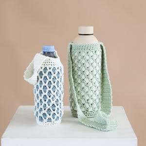 Peut inclure: Deux bouteilles d'eau dans des housses en crochet faites à la main. La bouteille de gauche a une housse blanche à motif en treillis avec une anse courte. La bouteille de droite est dans une housse en crochet vert clair avec une longue bandoulière.