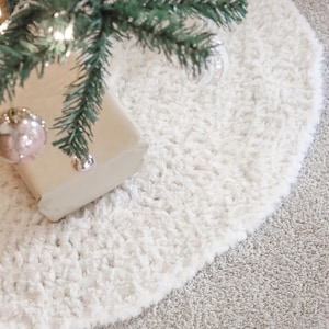 May include: A cream-colored, round Christmas tree skirt made of a soft, textured material. The skirt is placed under a green Christmas tree with ornaments and lights. The skirt has a fluffy, plush appearance.