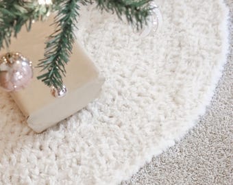 May include: A cream-colored, round Christmas tree skirt made of a soft, textured material. The skirt is placed under a green Christmas tree with ornaments and lights. The skirt has a fluffy, plush appearance.