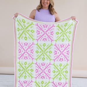 Puede incluir: Una manta de ganchillo hecha a mano con un patr&oacute;n geom&eacute;trico repetitivo en blanco, rosa y verde lima. La manta tiene un borde blanco con ribete rosa. El dise&ntilde;o incluye motivos florales y de mariposas.
