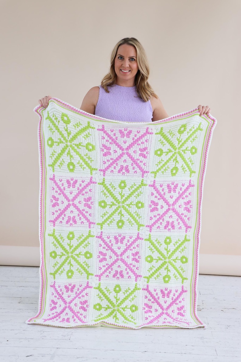 Puede incluir: Una manta de ganchillo hecha a mano con un patr&oacute;n geom&eacute;trico repetitivo en blanco, rosa y verde lima. La manta tiene un borde blanco con ribete rosa. El dise&ntilde;o incluye motivos florales y de mariposas.