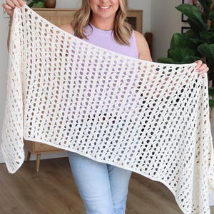 May include: Cream-colored, open-weave crochet shawl. The shawl is held up, showcasing its intricate pattern and texture. The handmade accessory is a light, airy design, perfect for layering. The color is a soft, off-white, suitable for various outfits.