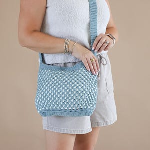 May include: A dusty blue crochet crossbody bag featuring a textured, open-weave diamond pattern on the main body, a solid flat base, and a wide, integrated shoulder strap. The bag is constructed from yarn with a matte finish and consistent stitching.