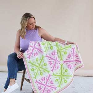 Puede incluir: Una manta de ganchillo blanca con un patr&oacute;n geom&eacute;trico en rosa y verde lima. La manta presenta un dise&ntilde;o de copo de nieve y un borde rosa y blanco. La manta se muestra sobre el regazo de una persona. La manta est&aacute; hecha de hilo suave.