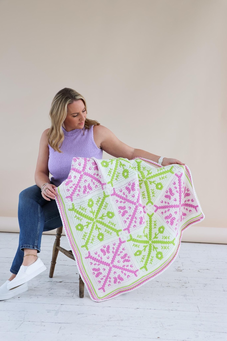 Puede incluir: Una manta de ganchillo blanca con un patr&oacute;n geom&eacute;trico en rosa y verde lima. La manta presenta un dise&ntilde;o de copo de nieve y un borde rosa y blanco. La manta se muestra sobre el regazo de una persona. La manta est&aacute; hecha de hilo suave.