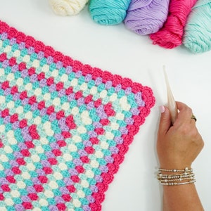 May include: A close-up of a crocheted blanket with a pink, white, blue, and purple color scheme. The blanket is being worked on with a crochet hook.