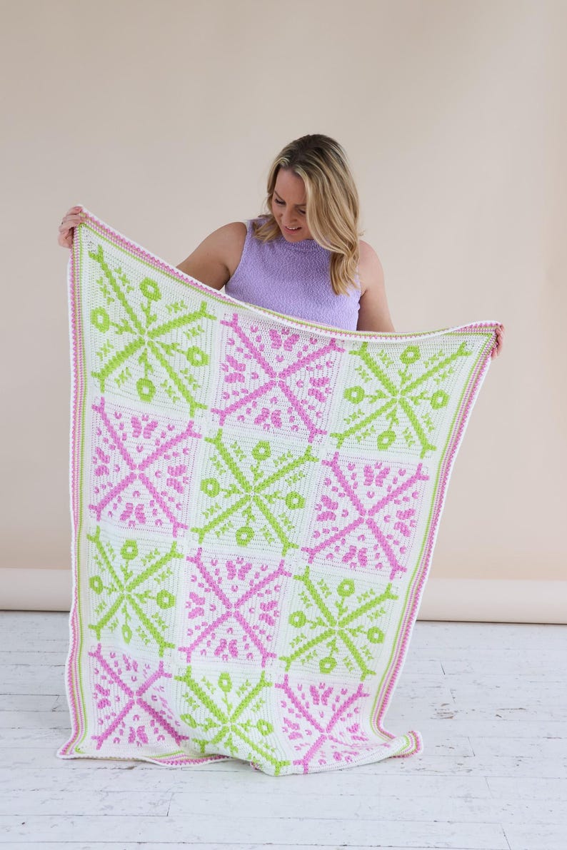 Puede incluir: Una manta de ganchillo hecha a mano con un patr&oacute;n geom&eacute;trico repetido en blanco, verde claro y rosa. La manta se sostiene, mostrando el intrincado dise&ntilde;o y la combinaci&oacute;n de colores. Los bordes est&aacute;n acabados con un borde rosa.