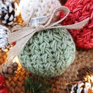 May include: Close-up of handmade Christmas ornaments. A light green, woven-style ornament is in focus, tied with a burlap bow. Other ornaments are visible in white and red. Pinecones and red berries are scattered around the ornaments.