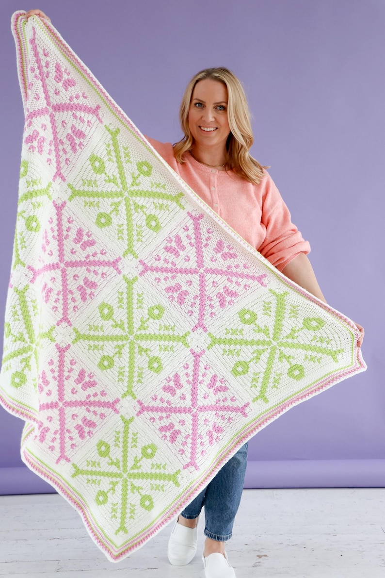 Puede incluir: Una manta de ganchillo hecha a mano con un patr&oacute;n geom&eacute;trico en blanco, rosa y verde. La manta presenta un dise&ntilde;o floral repetido y se sostiene sobre un fondo p&uacute;rpura. La manta es un art&iacute;culo de decoraci&oacute;n del hogar acogedor.
