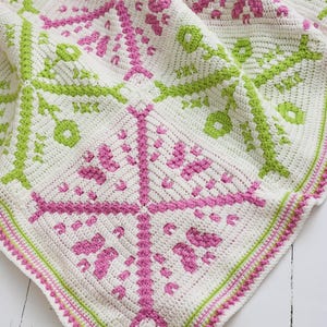 Puede incluir: Una manta de ganchillo hecha a mano con un patr&oacute;n cuadrado. La manta presenta una base blanca con dise&ntilde;os geom&eacute;tricos rosas y verdes. El borde est&aacute; acabado con un ribete rosa y verde, creando un textil acogedor y decorativo.