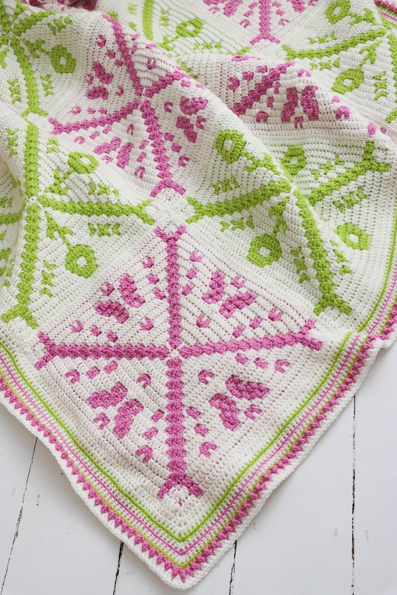 Puede incluir: Una manta de ganchillo hecha a mano con un patr&oacute;n cuadrado. La manta presenta una base blanca con dise&ntilde;os geom&eacute;tricos rosas y verdes. El borde est&aacute; acabado con un ribete rosa y verde, creando un textil acogedor y decorativo.