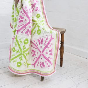 Puede incluir: Una manta blanca de ganchillo con un patr&oacute;n geom&eacute;trico en rosa y verde claro, sobre un taburete de madera. La manta presenta un dise&ntilde;o floral y un borde rosa y verde. El fondo es una pared y un suelo blancos.