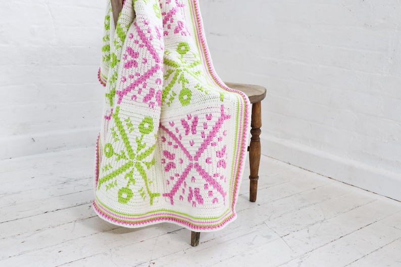 Puede incluir: Una manta blanca de ganchillo con un patr&oacute;n geom&eacute;trico en rosa y verde claro, sobre un taburete de madera. La manta presenta un dise&ntilde;o floral y un borde rosa y verde. El fondo es una pared y un suelo blancos.