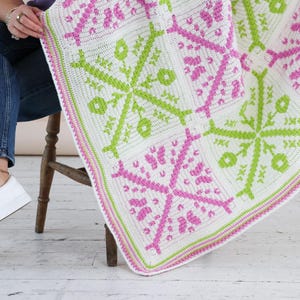 Puede incluir: Una manta de ganchillo blanca, rosa y verde con un patr&oacute;n floral geom&eacute;trico. La manta est&aacute; doblada sobre el regazo de una persona, mostrando el intrincado dise&ntilde;o y la combinaci&oacute;n de colores. Los bordes de la manta est&aacute;n acabados con un borde rosa y verde.