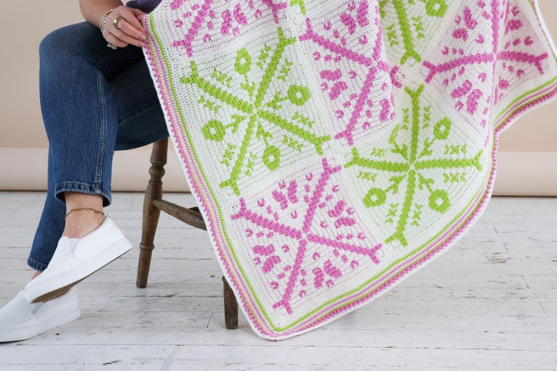 Puede incluir: Una manta de ganchillo blanca, rosa y verde con un patr&oacute;n floral geom&eacute;trico. La manta est&aacute; doblada sobre el regazo de una persona, mostrando el intrincado dise&ntilde;o y la combinaci&oacute;n de colores. Los bordes de la manta est&aacute;n acabados con un borde rosa y verde.