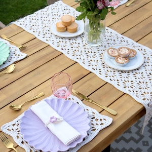 May include: A white crocheted table runner with a floral pattern, set on a wooden table with scalloped plates in light green and lavender, white napkins with floral rings, gold-toned cutlery, pink textured glasses, and small plates of food.