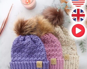 May include: Three crocheted beanies in purple, pink, and cream, each featuring a textured stitch pattern, a large faux fur pom-pom on top, and a small rectangular tan leather tag with the embossed letters BC attached to the folded brim.