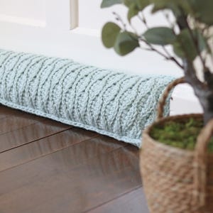 Op de afbeelding: Een lichtblauwe gehaakte deurstop met een gevlochten patroon. De deurstop ligt op een houten vloer naast een plant in een pot.