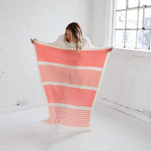 May include: A pink and white striped crocheted throw blanket. The blanket is being held up by a person wearing a white shirt.