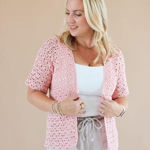 May include: A light pink crochet cardigan featuring a delicate floral-inspired openwork pattern, short sleeves, and a scalloped edge along the front opening and hem, worn over a plain white top and light-coloured shorts.