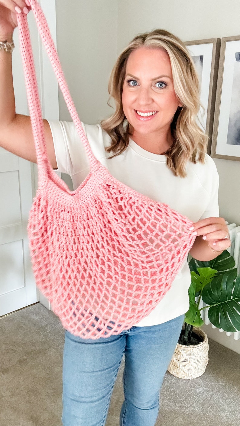 May include: A pink crocheted mesh bag with long straps. The bag is being held by a person wearing a white shirt and blue jeans.
