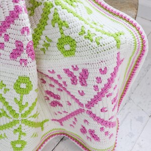 Puede incluir: Una manta blanca de ganchillo con un patr&oacute;n repetido en rosa y verde. La manta tiene un borde decorativo en rosa y verde. La manta est&aacute; doblada sobre una silla de madera.