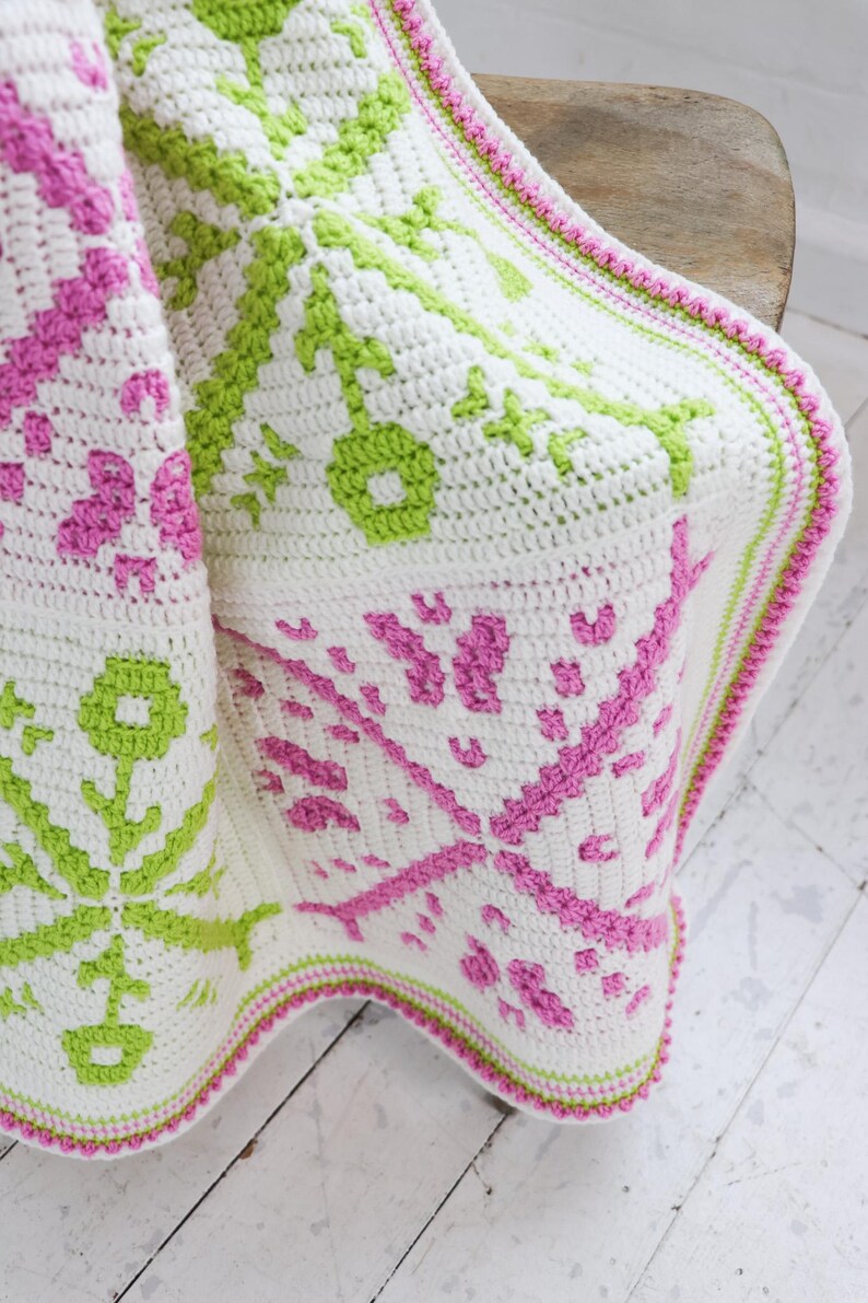 Puede incluir: Una manta blanca de ganchillo con un patr&oacute;n repetido en rosa y verde. La manta tiene un borde decorativo en rosa y verde. La manta est&aacute; doblada sobre una silla de madera.