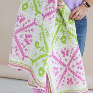Puede incluir: Una manta blanca de ganchillo con un patr&oacute;n repetido en rosa y verde. La manta presenta un dise&ntilde;o geom&eacute;trico con motivos florales y en forma de llave. Los bordes est&aacute;n adornados con rayas rosas y verdes. La manta est&aacute; sostenida por una persona.