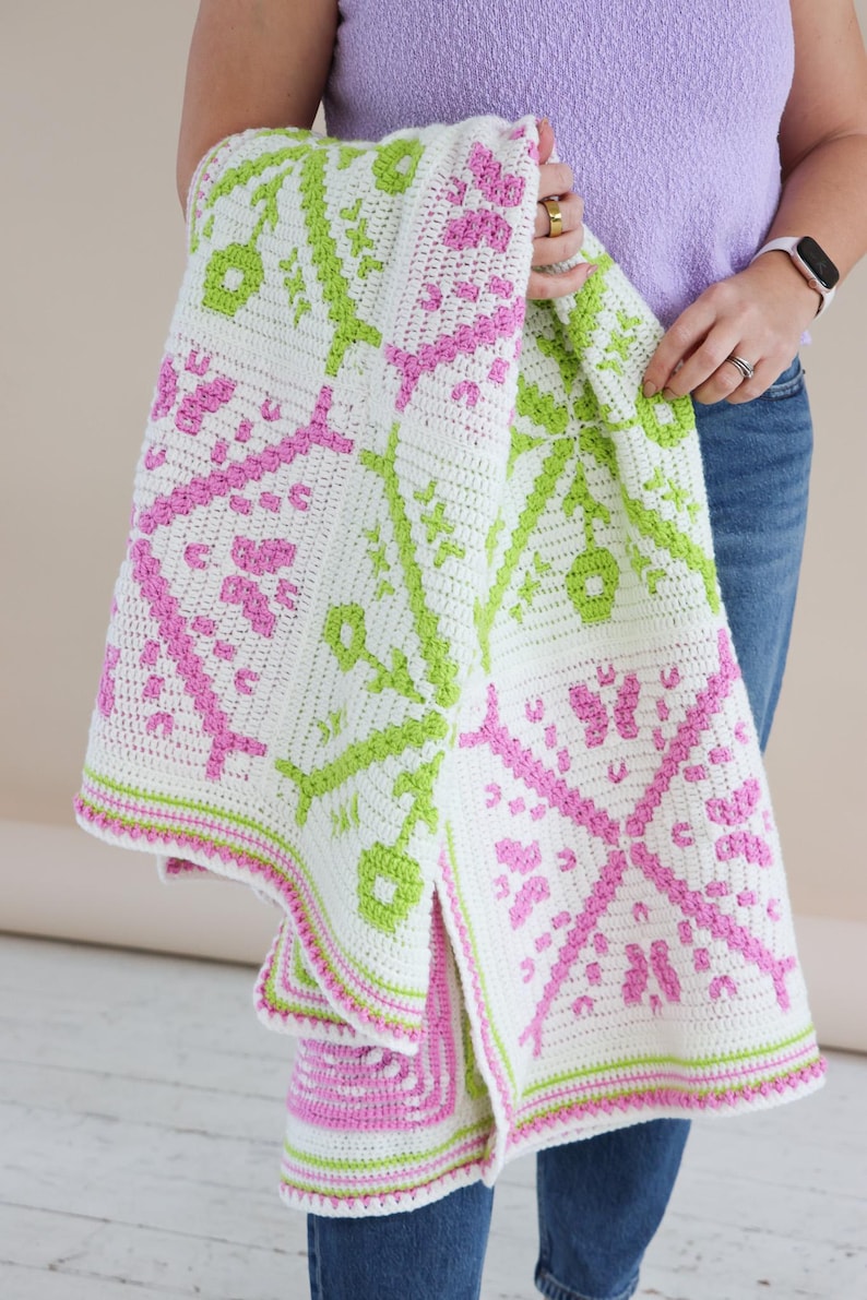 Puede incluir: Una manta blanca de ganchillo con un patr&oacute;n repetido en rosa y verde. La manta presenta un dise&ntilde;o geom&eacute;trico con motivos florales y en forma de llave. Los bordes est&aacute;n adornados con rayas rosas y verdes. La manta est&aacute; sostenida por una persona.