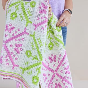 Puede incluir: Una manta blanca de ganchillo con un patr&oacute;n repetitivo de dise&ntilde;os florales y geom&eacute;tricos rosas y verdes. La manta se sostiene, mostrando los intrincados detalles del textil hecho a mano. Los bordes de la manta est&aacute;n acabados con un borde rosa y verde.