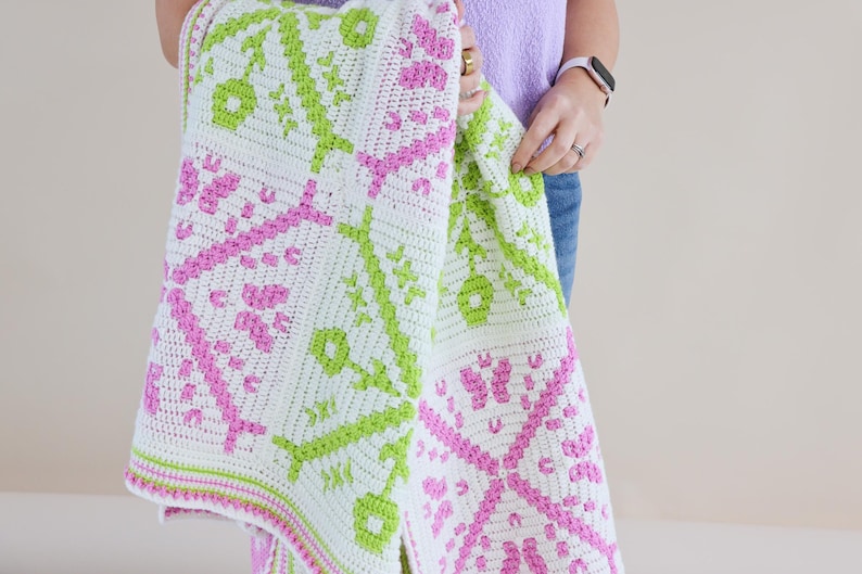 Puede incluir: Una manta blanca de ganchillo con un patr&oacute;n repetitivo de dise&ntilde;os florales y geom&eacute;tricos rosas y verdes. La manta se sostiene, mostrando los intrincados detalles del textil hecho a mano. Los bordes de la manta est&aacute;n acabados con un borde rosa y verde.