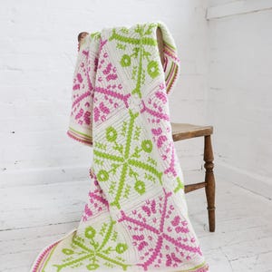 Puede incluir: Una manta blanca de ganchillo con dise&ntilde;os florales rosas y verde lima, sobre una silla de madera. La manta tiene un borde a rayas. La silla es de madera. Medidas aproximadas: 150 cm x 200 cm.