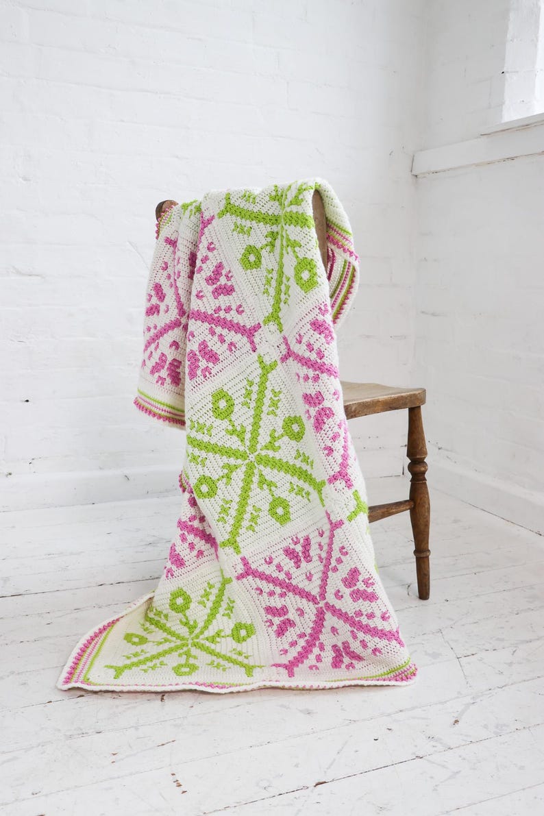 Puede incluir: Una manta blanca de ganchillo con dise&ntilde;os florales rosas y verde lima, sobre una silla de madera. La manta tiene un borde a rayas. La silla es de madera. Medidas aproximadas: 150 cm x 200 cm.