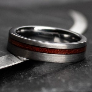 May include: A close-up of a silver ring with a dark reddish-brown wood inlay. The ring has a brushed metal finish and a sleek, modern design. The ring is resting on a dark, textured surface.