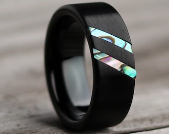 May include: A black ring with two diagonal inlays of iridescent abalone shell. The ring has a brushed metal finish and a polished interior. The abalone shell displays shades of blue, green, and pink.