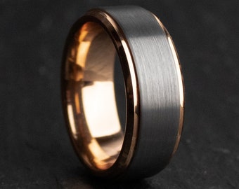 May include: A close-up of a wedding band with a brushed gray center and a rose gold interior and trim. The ring is set against a dark, textured background, highlighting its metallic sheen and design.