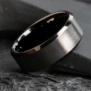 May include: A close-up of a black ring with a brushed metal finish. The ring has a polished silver edge and a dark interior. The ring is set against a dark, textured surface.