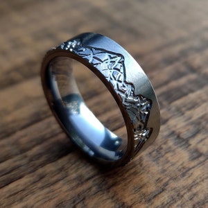 May include: A silver-colored ring with a mountain range design engraved around the band. The ring has a flat top and a polished finish, reflecting the light. The mountain range is detailed with peaks and valleys.
