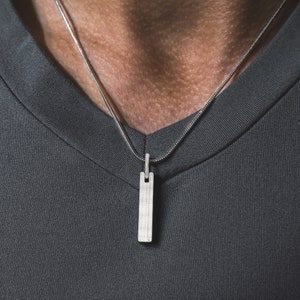 May include: A silver pendant necklace with a rectangular, brushed metal pendant. The pendant hangs from a delicate silver chain. The necklace is worn against a gray v-neck shirt.
