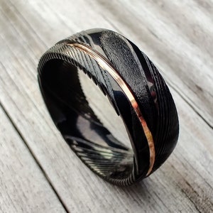 May include: A black ring with a textured surface and a thin, copper-colored stripe. The ring has a unique pattern and is set against a light wood grain background. The ring's design is eye-catching and modern.