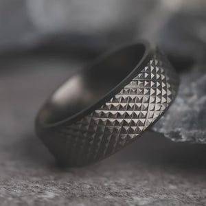 May include: A dark gray ring with a geometric, diamond-shaped pattern. The ring is made of a matte metal and has a smooth interior. The ring is a band style and appears to be a men's or unisex accessory.