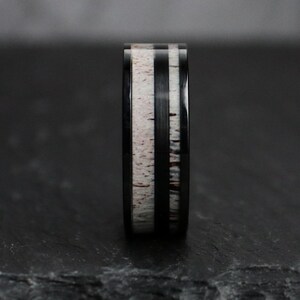 Puede incluir: Un anillo negro con una banda central de material moteado blanco y marr&oacute;n, flanqueado por dos bandas negras. El anillo tiene un acabado pulido, mostrando el contraste entre los materiales.