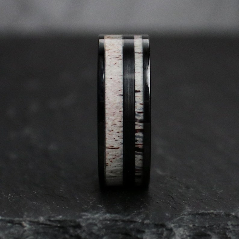Puede incluir: Un anillo negro con una banda central de material moteado blanco y marr&oacute;n, flanqueado por dos bandas negras. El anillo tiene un acabado pulido, mostrando el contraste entre los materiales.